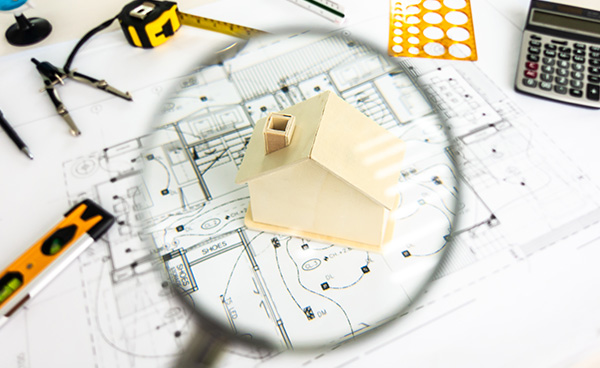 Sachverständiger Schall- und Wärmeschutz, eine Lupe gerichtet über ein Holzspielzeughaus auf einem Architektenplan symbolisch für Begutachtung